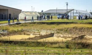 A group of participants walks along a gravel path from a pavilion toward a community garden during a conference. Greenhouses and a white van are visible in the background, along with an open field under a sunny sky. In the foreground, there is a grassy area with erosion fencing and a small pond.
