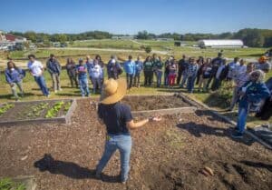 A woman wearing a wide-brimmed straw hat speaks to a group of participants gathered in a semi-circle around raised garden beds in a community garden. She gestures with her hands while presenting. The attendees are diverse in age and background, listening attentively under a bright sunny sky. The garden is surrounded by open fields, greenhouses, and other farm structures in the background.