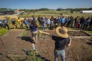 Morgan Malone, wearing a wide-brimmed straw hat, gestures toward a group of participants gathered around raised garden beds in a community garden. She speaks while the group listens attentively, standing on a grassy area under a clear blue sky. The garden features rows of plants, mulch, and fencing. In the background, a large building, vehicles, and open farmland are visible.