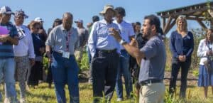 Biswanath Dari, wearing a gray shirt and holding a microphone, gestures while speaking to a group of participants in a Community Garden. The group listens attentively, with some individuals wearing hats, sunglasses, and name tags. A wooden pavilion and greenery are visible in the background under a sunny blue sky.