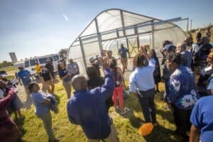 Trequan McGee, Ph.D., stands in front of a high tunnel structure during a presentation as part of the Regional Small Farms Conference. He addresses a group of participants, some of whom have raised hands to ask questions. The scene includes the high tunnel, a sunny blue sky, a van, and a distant silo in the background.