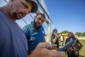 Trequan McGee, Ph.D., N.C. A&T State University Cooperative Extension Horticulture Extension Specialist, interacts with a farmer near a high tunnel, both looking at a mobile device. Other participants, including a woman holding a plant stalk, engage in conversation in the background under a clear blue sky.
