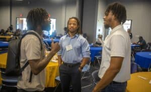 Three men standing in a group engage in conversation in a conference setting. The man on the left has dreadlocks and a backpack, the man in the center wears a blue button-up shirt with a name badge, and the man on the right wears a white polo shirt. The background shows round tables with yellow and blue tablecloths and other attendees seated in the room.
