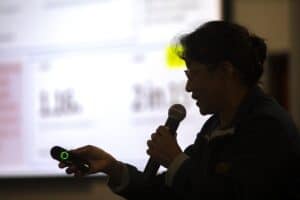A woman is speaking into a microphone while holding a presentation remote. She is silhouetted against a bright, blurred screen displaying text and graphics.