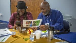 Pat Smith-Jones, wearing a black hat and glasses, smiles while looking at her phone, and Charles Jones, in a blue shirt, examines a sheet with images of plants. They are seated at a table covered with papers, a plate of fruit, and drinks during the Regional Small Farms Conference.