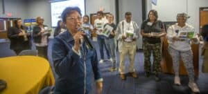 Kathleen Liang, Ph.D., speaks into a microphone while leading a group activity during her specialty crops presentation at the Regional Small Farms Conference. Behind her, participants stand in a line holding printed sheets with images of fruits and vegetables. A slide in the background reads, "Let Us Play a Game: Do You Know Your Specialty Fruit and Vegetables?"