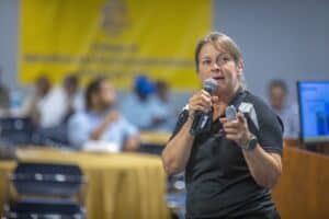 A woman in a black polo shirt speaks into a microphone while holding a presentation clicker. She gestures toward the audience, which includes seated individuals in a blurred background. Behind her is a yellow banner and a screen with an out-of-focus presentation slide.