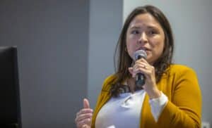 A woman wearing a mustard-yellow blazer speaks into a microphone during a presentation. She is standing in front of a neutral background, with part of a screen visible to the left.