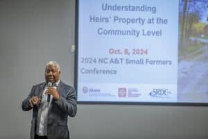 A man in a plaid blazer speaks into a microphone while gesturing during a presentation. Behind him, a slide is displayed on a large screen with the title "Understanding Heirs' Property at the Community Level" along with logos and conference details.