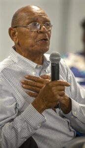 An elderly man wearing glasses and a striped button-up shirt holds a microphone in one hand while placing his other hand on his chest. He appears to be speaking, with an engaged and expressive facial expression. The background is softly blurred.