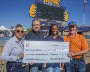 Four people standing in front of a large check awarded to Nina Story for $2,500, with a sports stadium in the background. The check is labeled as an 'AG Day Scholarship' and is signed by the CAES Advisory Board. The group is smiling, and a scoreboard at Truist Stadium is visible above them, showing a football game in progress.