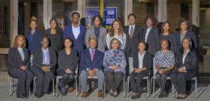alt tag: A group of 17 professionally dressed individuals pose for a group photo in two rows outside a brick building with large windows. The front row consists of five women and two men sitting on chairs, while the back row has ten standing individuals. The group is diverse in gender and ethnicity, all smiling warmly at the camera.