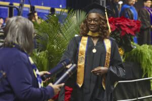 A graduate in a black cap and gown with a gold stole labeled "NC A&T" receives her diploma during the commencement ceremony. She smiles as she reaches for the diploma, with festive greenery and poinsettias in the background.