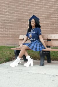 A young woman sits on a wooden bench outdoors, wearing a blue graduation cap, a navy 