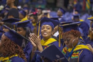 A graduate in a blue cap and gown smiles and claps while seated during a commencement ceremony. Surrounding graduates, some wearing stoles with colorful patterns, are reading programs or watching the event in the indoor arena.