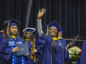 A graduate in a blue cap and gown smiles and waves while holding a diploma cover during a commencement ceremony. Other graduates in similar attire, some wearing stoles, stand in the background near a stage with dark curtains.