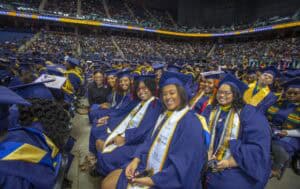 A group of smiling graduates in blue caps and gowns seated together in rows during a commencement ceremony in an indoor arena. Some graduates wear stoles and cords, and decorated caps are visible. A large audience fills the background, with banners and signage congratulating the graduates.