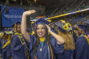 A smiling graduate wearing a blue cap decorated with a butterfly and yellow flowers waves enthusiastically during a commencement ceremony in an indoor arena. Other graduates in blue caps and gowns are visible in the background, along with a stage and audience seating.