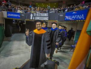 A faculty member in academic regalia smiles and flashes a peace sign while walking in a processional during a commencement ceremony. Other faculty members follow behind, and the audience watches from the stands.