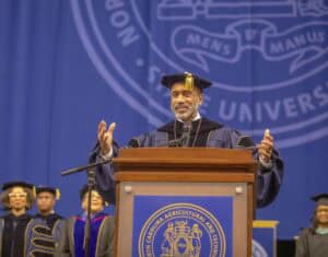 A distinguished speaker in academic regalia stands at a podium delivering a speech during a commencement ceremony. Behind him, a large blue university banner displays the seal of North Carolina Agricultural and Technical State University.