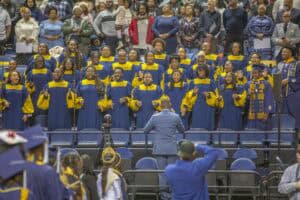 A choir dressed in blue and gold robes performs during a commencement ceremony, led by a conductor in a blue suit. Audience members watch and stand in the background.