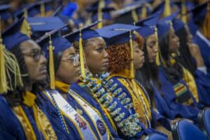 Graduates in blue and gold regalia sit attentively during a commencement ceremony. One graduate in the center wears a necklace of dollar bills, while others focus forward.
