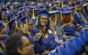 Graduates in blue and gold regalia sit in rows during a commencement ceremony. A smiling graduate with glasses waves to the camera while others adjust their caps or look forward.