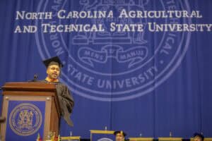 A commencement speaker in a black cap and gown stands at a podium delivering a speech. Behind him, a large blue banner displays the name and seal of North Carolina Agricultural and Technical State University.