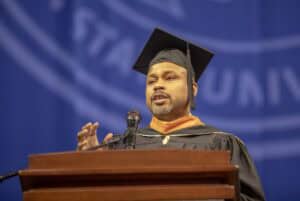 A commencement speaker in a black cap and gown delivers a speech at a podium, gesturing with one hand. A large blue university banner is visible in the background.