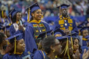 A graduate in blue and gold regalia stands smiling with pride during a commencement ceremony, wearing a colorful Kente stole. Other graduates in caps and gowns surround her, seated and applauding.