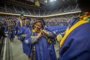 A group of graduates in blue and gold regalia smile and celebrate while walking into a large arena during a commencement ceremony. The central graduate enthusiastically flashes a hand sign while surrounded by peers.