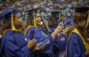 College graduates in blue caps and gowns take photos and hold diplomas during a commencement ceremony. The middle graduate smiles as she takes a selfie with her phone, holding her diploma in the other hand.