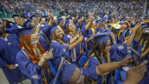 A large group of college graduates wearing blue caps and gowns celebrate together at a graduation ceremony in an indoor arena. The graduates are smiling, holding up phones, and cheering, with a diverse crowd seated in the background.