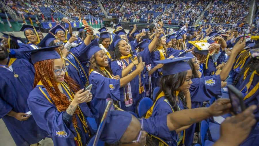 A large group of college graduates wearing blue caps and gowns celebrate together at a graduation ceremony in an indoor arena. The graduates are smiling, holding up phones, and cheering, with a diverse crowd seated in the background.