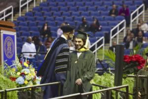 A graduate in a green cap and gown walks across the stage during the NC A&T commencement ceremony, smiling as he is greeted by a university official in formal academic regalia. The stage is decorated with flowers, greenery, and a university seal backdrop.