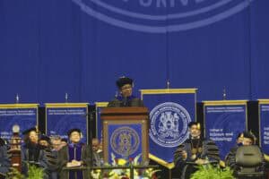 A keynote speaker in academic regalia stands at a podium during the NC A&T commencement ceremony. Behind her are large blue and gold banners with the university's seal and college names, while faculty members sit on stage in formal regalia.