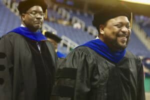 Two faculty members in black academic regalia with blue velvet hoods stand during a commencement ceremony. The faculty member in the foreground smiles brightly, while the one in the background looks forward.