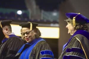 Two faculty members in academic regalia stand on stage during a commencement ceremony. Both wear black robes with blue velvet accents and caps with gold tassels, smiling as they look toward the audience.