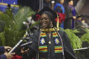 A graduate student in a black cap and gown smiles while receiving a diploma. She wears a colorful Kente cloth stole embroidered with the NC A&T logo and honor cords, symbolizing her achievements.
