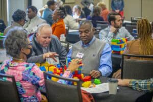 A group of adults at tables in a conference room participate in an activity building colorful block structures, engaging in conversation and collaboration. Coffee cups and papers are also on the tables.