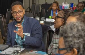 A man in a blazer gesturing while engaged in conversation with women at a conference.