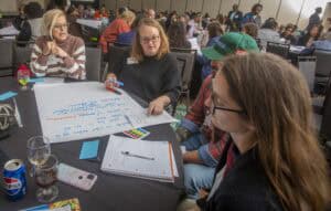A group collaborates at a table, discussing ideas and taking notes on large paper in a conference setting.