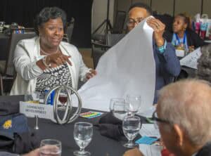 A woman in a white blazer gestures and speaks animatedly during a group discussion at a conference table. Next to her, a man in glasses and a blue blazer holds up a large blank sheet of paper. The table is labeled “Community Development” and is scattered with water glasses, papers, and markers. Other attendees are visible in the background, engaged in their discussions.
