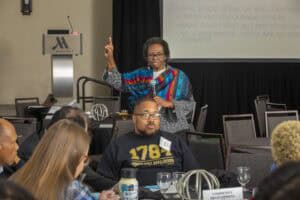 A woman, identified as Yolanda Burwell, Ph.D., stands at the front of a conference room, holding a microphone and raising her hand while speaking to attendees. She is wearing a vibrant multicolored shawl over a black and white outfit. In the foreground, participants sit at round tables, listening attentively. A large screen behind her displays session instructions such as "Give voice, speak up, and contribute" and "No judgments - there are no right answers."