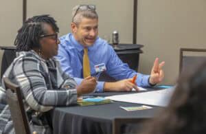 A man wearing a blue shirt and gold tie, identified as Mark Light, Ph.D., consults with a teammate during a group discussion at a conference table. He gestures with two fingers while holding an orange marker. Beside him, a woman in a plaid jacket listens attentively, holding a pen and a notebook. The table is covered with a black tablecloth, a large sheet of paper, documents, and other materials for the session.