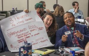 Two individuals, a woman with long curly hair holding a large handwritten poster and a man in a blue blazer pointing playfully, are engaged in a lively discussion at a conference. The poster outlines “3 Priorities” for agriculture and food systems, including topics like “Farmland Loss” and “Food Access.” The table in front of them contains documents, a notebook, beverages, and a reusable cup, indicating active participation.