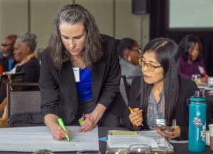 Two women work together at a table during a conference session. The woman on the left, with long gray-streaked hair, is writing with a green marker on a large sheet of paper. The woman on the right, wearing glasses and a black blazer, holds a pen and a smartphone while reviewing documents on the table. A teal water bottle and various papers are scattered across the table, and other conference attendees are visible in the background.