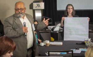 A man with glasses, a beard, and a gray suit holds a microphone and gestures with his hand while speaking during a conference session. Behind him, a woman with long hair and a black outfit holds up a large white poster with handwritten green notes titled "Nutrition," listing points such as "Nutrition-rich food," "Education," "Nutrient Value," and "Food Safety." A table with papers, cups, and a water bottle is visible in the foreground.