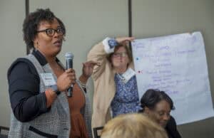 A woman in glasses and a plaid blazer, identified as Shannon Wiley, Ph.D., speaks into a microphone while gesturing with her hand during a conference session. Behind her, another woman in a beige jacket holds up a large white poster with handwritten notes in blue and purple marker, including the heading "Family Wellbeing" and subtopics like "Chronic Disease Prevention" and "Managing Finances." A seated woman listens intently in the foreground.