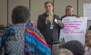 A man in a black suit and yellow lanyard speaks into a microphone while holding a name badge, standing in front of a seated audience. Behind him, another man in glasses holds a large white poster with handwritten notes in pink marker, including "Priorities" and points like "Diversity of all representations" and "Building partnerships & relations." A woman in colorful, patterned attire is seen from the back, listening attentively.